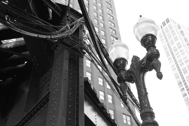 Riveted steel ironwork of an elevated train structure alongside an ornate double-globe cast-iron lamppost, with skyscrapers behind